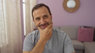 Pembe duvarları ve gri mobilyalarıyla oturma odasında oturan düşünceli, orta yaşlı bir adam..