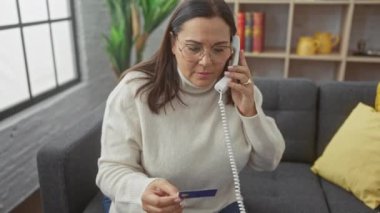 Orta yaşlı bir kadın evinin oturma odasında elinde kredi kartıyla ev telefonuyla konuşuyor.