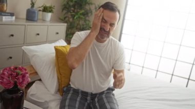 Orta yaşlı bir adam baş ağrısı çekiyor. Yatak odasında pijamalarıyla oturuyor.