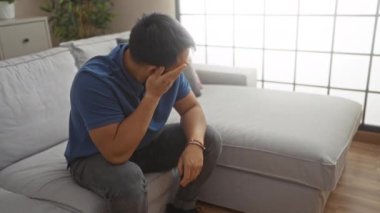 Genç yakışıklı Çinli bir adam oturma odasında bir kanepede oturuyor, düşünceli ve düşünceli bir şekilde, sıcak evine bakıyor..