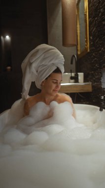 Huzurlu bir kadın, sofistike bir otel banyosunda lüks bir köpük banyosunun keyfini çıkarıyor, rahatlama ve kendine bakmanın vücut bulmuş hali..
