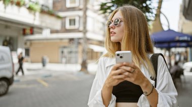 Güneşli şehir caddesinde güneş gözlüklü sarışın kadın akıllı telefon kullanıyor..