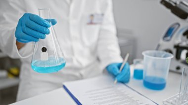 Laboratuvarda bir bilimadamı elinde mavi sıvıyla beyaz önlük ve koruyucu eldiven giyerek notlar alan bir şişe tutuyor..
