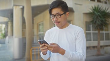 Beyaz gömlekli genç Asyalı bir adam güneşli bir şehir caddesinde akıllı telefonunu kullanıyor..