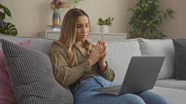 Evde dizüstü bilgisayarda çalışan sarışın bir kadın, şehir merkezindeki bir dairede koltukta oturuyor, arka planda bitkiler ve rahat bir dekor var..