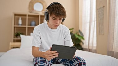 Kafkasyalı genç bir çocuk yatak odasında otururken bir tablete odaklanmış, kulaklık ve pijama giyiyordu..