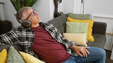 Gri saçlı, rahat bir orta yaşlı adam kulaklıkla müzik dinlemekten zevk alır, rahat bir oturma odasında renkli bir kanepeye uzanır..