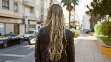 Deri ceketli sarışın bir kadının şehrin kaldırımında dikilirkenki görüntüsü..