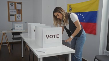 Genç, sarışın, çekici bir kadın, kapalı Venezuela seçim odasında arka planda Venezuela bayrağıyla oy vermeye katılıyor..