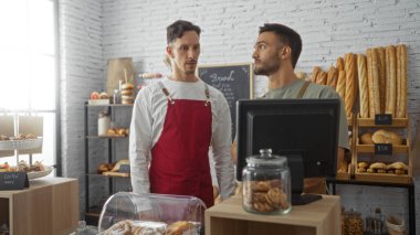 İki adam bir pastanede birlikte çalışıyor, çeşitli ekmek ve hamur işleri ile birlikte serginin önünde duruyorlar, biri kırmızı önlük takıyor ve diğeri tezgahın üstündeki bir şeyi işaret ediyor.