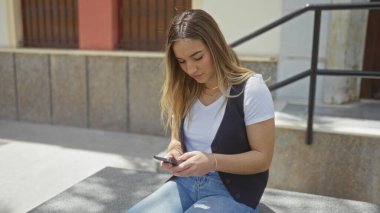 Genç, çekici sarışın bir kadın şehir caddesinde oturuyor, akıllı telefonuna odaklanmış..