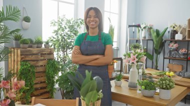 Güvenilir Afro-Amerikalı kadın kollarını kavuşturmuş yeşil bir çiçekçi dükkanında dikiliyor bitkilerle çevrili.
