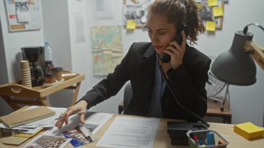 Odaklanmış bir kadın dedektif telefonda konuşurken karmaşık polis masasında kanıtları analiz eder..
