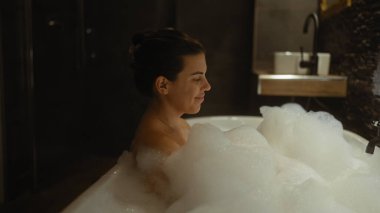 Genç İspanyol kadın loş bir ev banyosunda rahatlatıcı köpük banyosunun keyfini çıkarıyor.