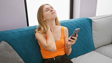 Sarışın bir kadın, kapalı bir kanepede akıllı bir telefona bakarken boynunu tutmaktan rahatsızlık duyuyor..