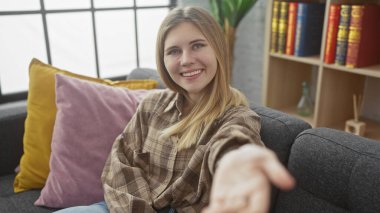 Genç sarışın bir kadın gülümsüyor, elini uzatıyor, rahat bir oturma odasında renkli yastıklar ve bir kitaplıkla oturuyor..