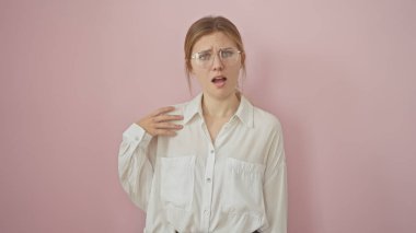 Gözlüklü kafası karışmış genç bir kadın pembe bir arka plana karşı duruyor ve kafa karışıklığını ifade ediyor..