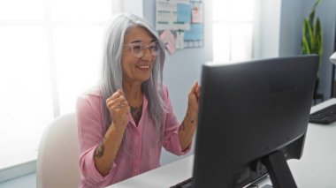 Ofis masasındaki başarıyı bilgisayar ve evrak işleriyle kutlayan kadın, gri saçlı, gözlüklü ve pembe gömlekli, pencere ışığı ve bitkilerle dolu, neşeli ve olgun