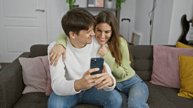 Bir adam ve bir kadın, bir kanepede akıllı telefon kullanarak, rahat bir oturma odasında birbirlerine sarıldılar..