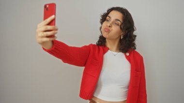 Güzel, genç, İspanyol, esmer bir kadın izole edilmiş beyaz bir arka planda kırmızı bir telefonla neşeli bir selfie çekiyor..