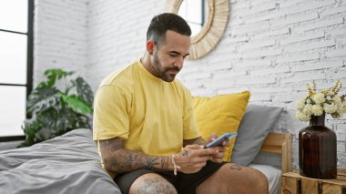 Gündelik kıyafetli İspanyol dövmeli bir adam modern bir yatak odası ortamında beyaz tuğlalı bir duvar kullanıyor..