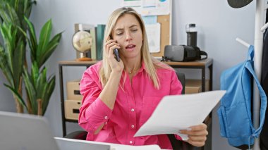 Pembe tişörtlü profesyonel sarışın kadın ofis ortamında birden fazla iş yapıyor, telefonda konuşuyor ve belgeleri okuyor..