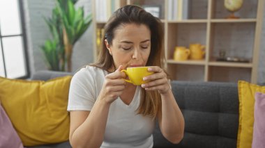 İspanyol kadın, modern oturma odasındaki canlı sarı aksanlı rahat bir koltukta kahvesini yudumluyor..