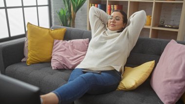 Rahat, yetişkin bir İspanyol kadın güzel dekore edilmiş bir oturma odasındaki renkli yastıklarla çevrili rahat bir kanepede vakit geçirir..