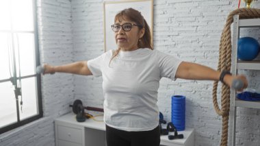 Olgun bir İspanyol kadın spor salonunda egzersiz yapıyor, iyi donanımlı bir odada odaklanmış bir ifadeyle halterleri kaldırıyor.