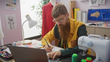 Sakallı kızıl saçlı bir adam iyi donanımlı bir terzi dükkanında tasarımlar çiziyor. Yaratıcılık ve moda uzmanlığını ima ediyor..