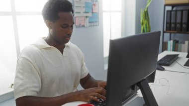 Ofiste çalışan genç bir adam, bilgisayarda yazı yazan, beyaz gömlek giyen, odaklanmış ve profesyonel, modern bir iş yerinde doğal ışık ve düzenli bir çevreye sahip.