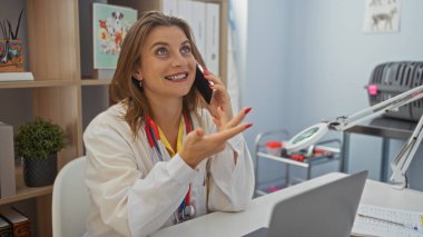 Klinikte gülümseyen kadın veteriner telefonda steteskopla konuşuyor. Çalışma masasında, parlak ve düzenli veterinerlik ortamı..
