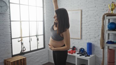 Genç, Latin kökenli hamile bir kadın spor salonunda kolunu yukarı doğru uzatıyor ve kapalı bir spor merkezinde annelik ve zindelik gösterisi yapıyor..