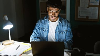 Yakışıklı, sakallı, ispanyol bir adam gece yarısı elinde dizüstü bilgisayar ve lambayla bürosundaki işine konsantre oluyor..