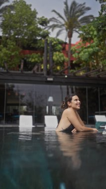 Genç bir kadın, tropik yeşilliklerle çevrili bir tatil beldesinin açık havuzunda huzurun tadını çıkarıyor..