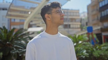 Şehir parkında oturan, gökyüzüne bakan, gözlüklü, beyaz gömlekli, şehir binaları ve yeşillikleri olan genç bir İspanyol..