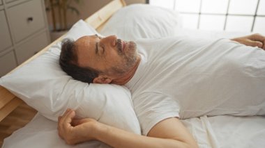 Beyaz tişörtlü İspanyol bir adam. Parlak yatak odasında, yüzünde barışçıl bir ifadeyle yatıyor.