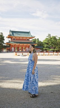 Beautiful hispanic woman confidently standing, enjoying and smiling while looking around the traditional heian jingu shrine, kyoto, japan clipart