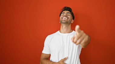 Gülümseyen genç İspanyol adam kırmızı arka planlı kamerayı işaret ediyor..