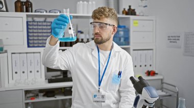 Genç, yakışıklı, sakallı, laboratuvar önlüklü beyaz bir adam laboratuarın içinde mavi bir sıvıyı inceliyor..