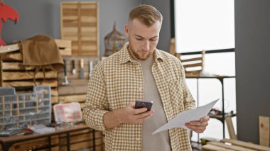 Akıllı telefon kullanan ve marangozluk atölyesinde evrak tutan sakallı yakışıklı bir genç..