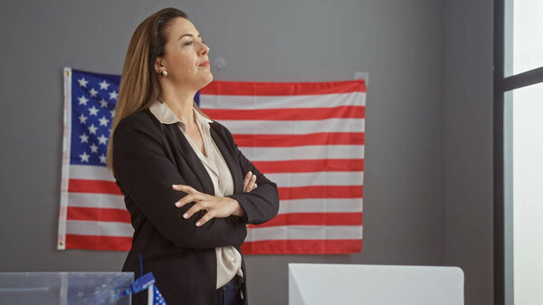 Уверенная латиноамериканка со скрещенными руками в избирательном центре колледжа, американский флаг на заднем плане.