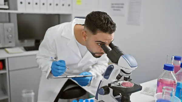 stock image A young man with a beard examines samples under a microscope in a laboratory setting.