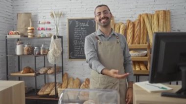 Önlüklü, gözlüklü ve bıyıklı genç adam büyüleyici bir dükkanda gülümsüyor ve pastane ürünlerini sergiliyor.