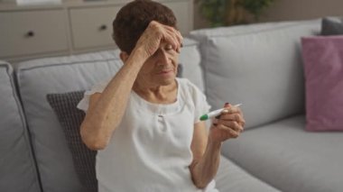 Oturma odasındaki gri bir koltukta oturan yaşlı bir kadın. Ateşini dijital termometreyle ölçüyor. Alnındaki bir elle ilgili görünüyor..