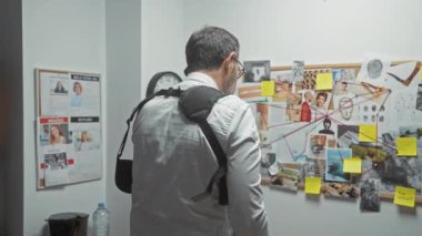 Sakallı, orta yaşlı bir adam, bir ofis ortamında cinayet soruşturması kurulunu inceliyor, akıllı bir telefonla konuşurken kanıtları işaret ediyor..
