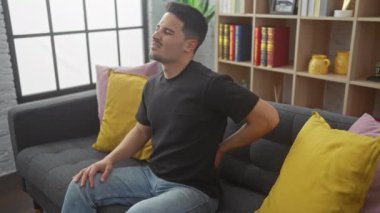 Yakışıklı, sakallı ve sakallı bir adam. Gri koltukta oturuyor. Evin içine doğru dokunuyor..