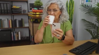 Gri saçlı, kahve içen ve modern ofis içinde kitaplık ve bitkilerle akıllı telefon kullanan olgun bir kadın.