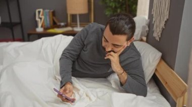 Bıyıklı genç İspanyol adam yatakta uzanıyor ve rahat yatak odasında akıllı telefon kullanıyor.