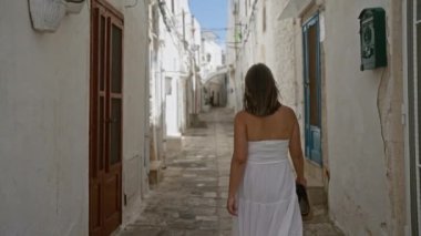 Genç bir İspanyol kadın, Puglia, İtalya, Avrupa 'daki eski Ostuni kasabasında dar ve tarihi bir sokakta elinde bir kamış çanta ve beyaz bir elbise ile yürüyor..